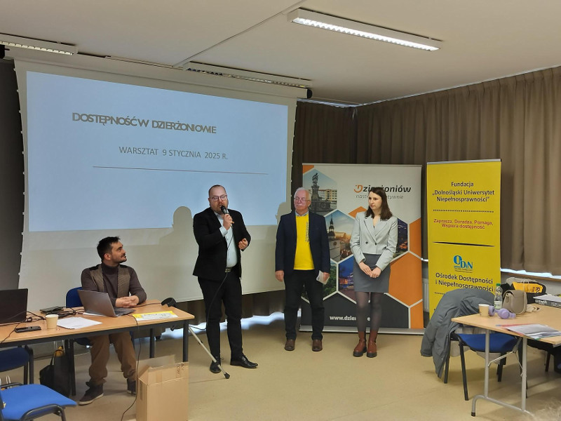 Prelegenci warsztatów w Dzierżoniowie. Stoją przed ekranem z tytułem spotkania. Obok stół, leżą na nim materiały. W tle stoją roll-upy organizatorów wydarzenia.