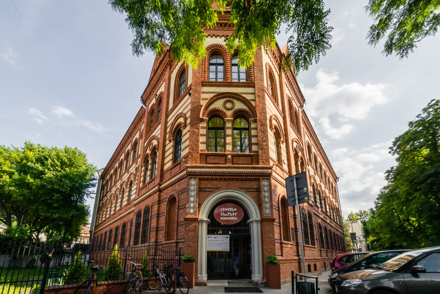na środku widoczny narożny budynek z czerwono kremowej cegły. Centralnie umieszczone drzwi wejściowe z napisem "Centrum Kultury Podgórza". Po bokach budynku zielone drzewa i kilka zaparkowanych samochodów