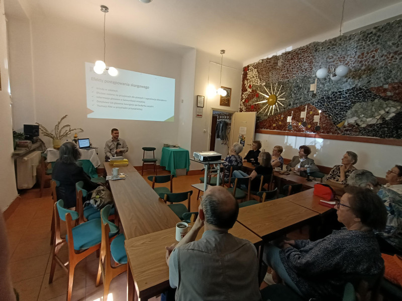 W jasnej sali z mozaiką na ścianie, prelegent stoi obok ekranu projekcyjnego. Prowadzi prezentację dla grupy słuchaczy, którzy siedzą przy połączonych stołach w kształcie litery U.