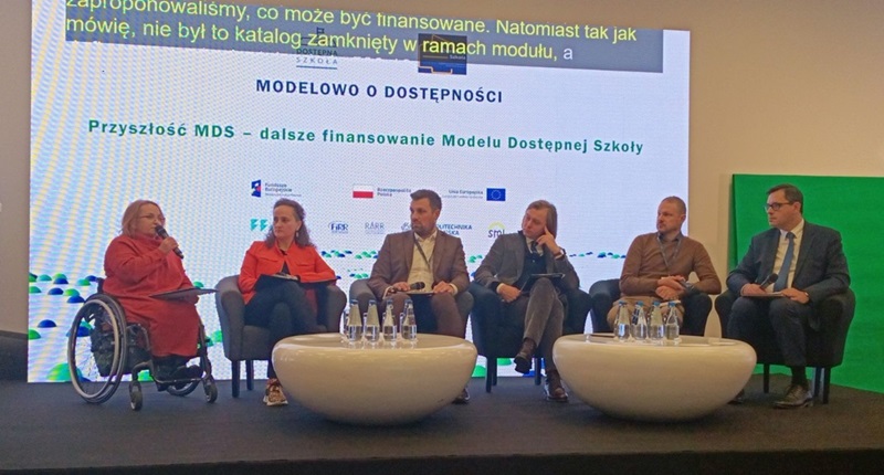 6 osób siedzi przy małych stolikach podczas rozmowy. Małgorzata Radziszewska z PFRON prezentuje temat. W tle plansza informacyjna z tytułem konferencji i panelu: „Modelowo o dostępności. Przyszłość MDS – dalsze finansowanie Modelu Dostępnej Szkoły”.